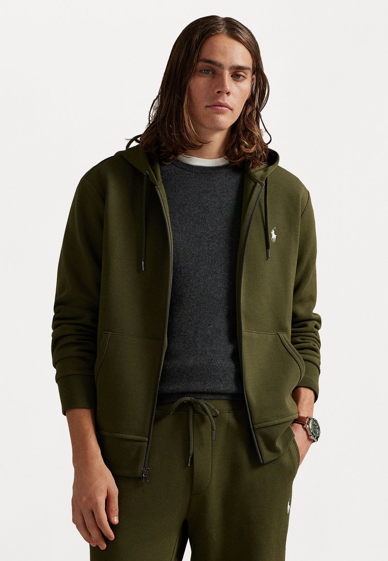 Ελαιόχρωμο φούτερ με φερμουάρ και κορδόνια, με δύο μπροσ�τινές τσέπες, συνδυασμένο με ένα γκρι πουλόβερ από κάτω. Περιέχει ένα μικρό λογότυπο στο στήθος.