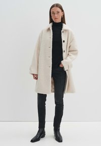 Manteau en laine texturé couleur crème, avec un large col, des boutons sur le devant et des poches latérales, porté sur un col roulé noir côtelé et un jean foncé.