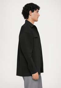 Fekete, gombos ing, álló gallérral és egyetlen mellzsebbel, sima, szőtt anyagból készült. Slim-fit szabás.