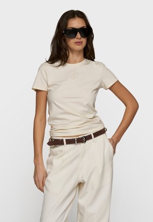 Donna con occhiali da sole neri oversize, camicia beige a maniche corte, pantaloni beige a vita alta e cintura marrone, che posa con una mano sui fianchi.