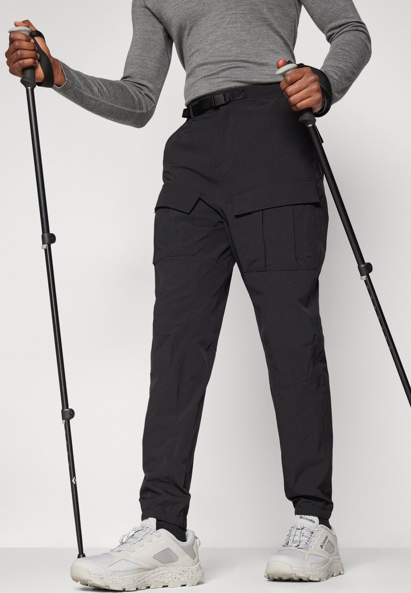 Czarne spodnie cargo z wieloma kieszeniami, wykonane z lekkiego materiału, z elastycznym mankietem na kostkach. W połączeniu z jasnoszarymi trampkami i kijkami trekkingowymi.