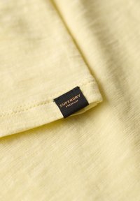 Tela de color amarillo claro con acabado texturizado, que presenta una etiqueta negra con la inscripción "SUPERDRY" en letras de color cobre cerca del borde.