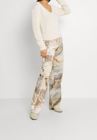 Pull blanc moelleux avec un col en V et des manches longues. Pantalons large à motifs multicolores avec des teintes terreuses et des designs abstraits. Chaussures beiges.