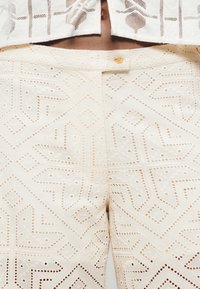 Pantalon couleur crème présentant des motifs géométriques découpés complexes, une fermeture éclair et un bouton, ainsi qu'un tissu à la texture lisse.