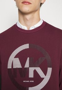 Sudadera burdeos con un logo redondo que presenta "MK" en rayas negras y blancas, y un cuello de barco sobre una camisa con cuello.