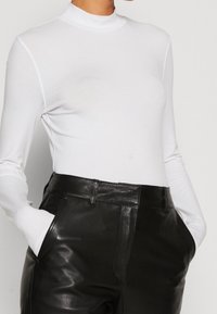 Weißes Langarm-Turtleneck-Oberteil aus dehnbarem Material, kombiniert mit hoch taillierten schwarzen Lederhosen mit sichtbaren Seitentaschen.