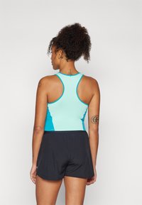 En tettsittende sportstopp i lys blå med racerback-design, kombinert med svarte shorts. Stoffer ser glatte ut, og det er synlige tatoveringer på armen.