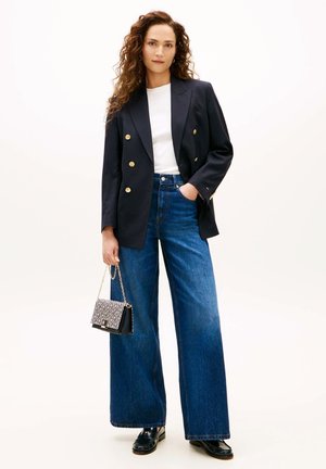 Femme aux cheveux bouclés portant un blazer noir croisé, un haut blanc, un jean bleu à jambes larges, des mocassins noirs, tenant un petit sac à main à motifs.