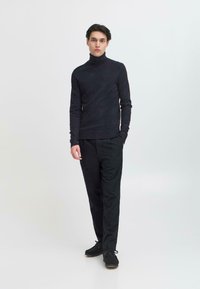 Dunkler Rollkragenpullover kombiniert mit schwarzen Hosen. Der Pullover hat eine glatte Textur, eine schmale Passform und lange Ärmel. Schwarze Schuhe runden das Outfit ab.