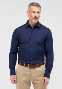 Mørkeblå, tætsiddende skjorte med lange ærmer og en tekstureret finish, knap-ned krave og matchende knapper. Kombineret med lyse beige bukser og et brunt bælte.