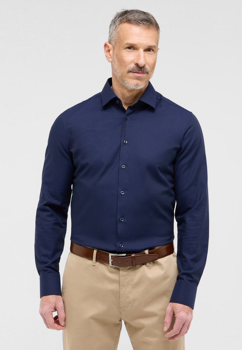 Marineblaues, tailliertes Langarmhemd mit strukturiertem Finish, mit Button-Down-Kragen und passenden Knöpfen. Kombiniert mit hellbeigen Hosen und einem braunen Gürtel.