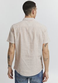Chemise à manches courtes en beige clair avec des rayures verticales blanches, dotée d'un col classique et d'un ourlet arrondi. Tissu texturé avec des coutures visibles.