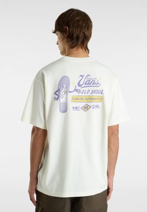 Creme kortærmet t-shirt med grafisk print på ryggen, der har lilla tekst og en skoillustration, lavet af blødt bomuldsstof.