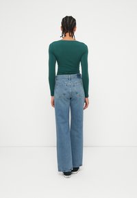 Μακρυμάνικο, εφαρμοστό πράσινο μπλουζάκι συνδυασμένο με ψηλόμεσα, φαρδιά ανοιχτού μπλε τζιν παντελόνια. Τα τζιν διαθέτουν πίσω τσέπες και κλασική υφή τζιν.