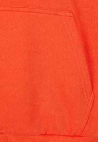 Oranžinės spalvos džemperio audinys su tekstūruotu paviršiumi. Yra didelė priekinė kišenė su sustiprintomis siūlėmis ir gumbuotais kraštais.