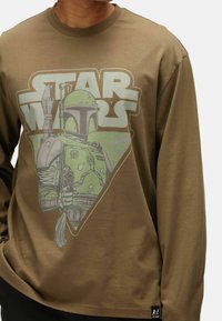 Camicia a maniche lunghe verde oliva con una stampa grafica di un personaggio in armatura e la scritta "STAR WARS" in lettere grasse.