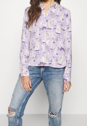 Femme portant une chemise lavande boutonnée avec des imprimés de statues classiques et un jean bleu clair déchiré.