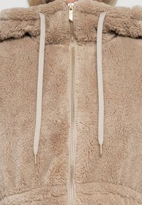 Sweat à capuche zippé en polaire beige avec une surface texturée, doté d'une capuche doublée, de cordons de serrage et de détails en zip doré.