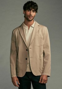 Blazer en coton beige avec revers crantés, deux poches avant, fermeture à un bouton, superposé sur un polo de couleur claire.