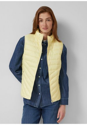 Młoda kobieta z długimi rudymi włosami, ubrana w jasnofioletową pikowaną kamizelkę na ciemnoniebieskiej dżinsowej koszuli i dżinsach, stoi na tle jednolitej ściany.