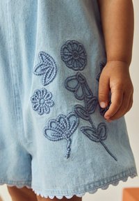 Ljusblå denimromper med blå broderade blommönster, scallopad fåll och en synlig hand som håller tyget.