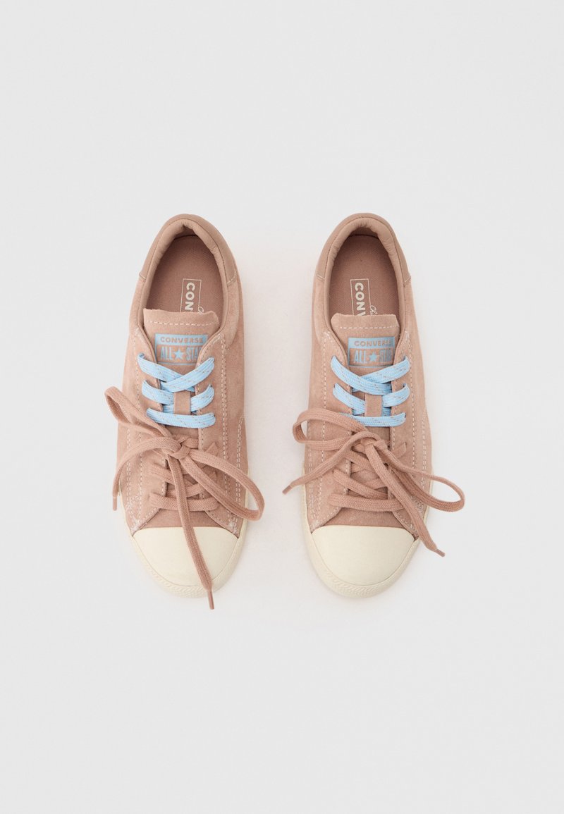 Paar beige Wildleder-Converse-Sneaker mit weißen Zehenkappen und unterschiedlichen Schnürsenkeln: rechter Schuh mit beigen Schnürsenkeln, linker Schuh mit blauen Schnürsenkeln.