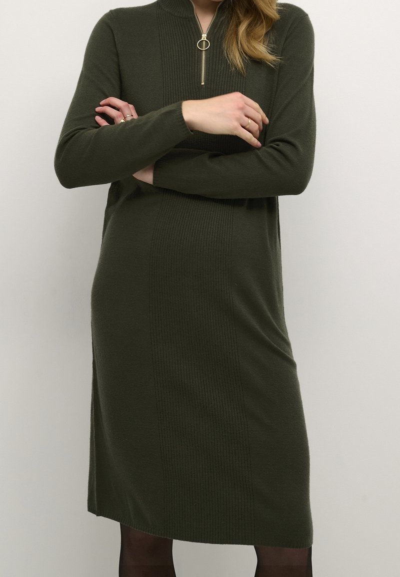Femme portant une robe en maille vert foncé à manches longues avec un col demi-fermeture éclair, les bras croisés, sur un fond clair.