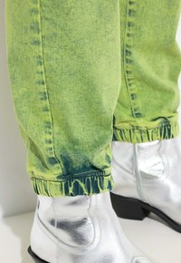 Joggers de mezclilla verde neón con puños elásticos, combinados con botas de vaquero plateadas brillantes que cuentan con detalles de costura y puntas afiladas.