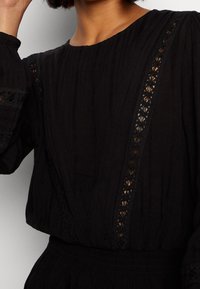 Mustat puserot, jotka on valmistettu teksturoidusta kankaasta, pyöreä pääntie, pitkät hihat ja monimutkainen pitsikoristelu edessä.