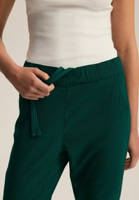 Zielone spodnie na gumce z regulowanym sznurkiem i bocznymi kieszeniami. Tkanina ma gładką fakturę i jest dopasowana w okolicy talii.