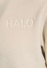 Gräddfärgad fleeceplagg med en texturerad yta, som har en präglad "HALO"-logotyp på framsidan. Inga andra synliga detaljer.