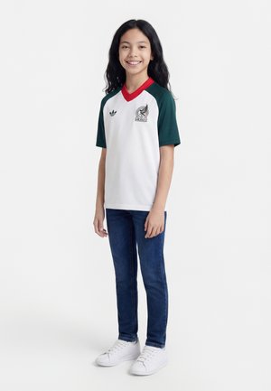 Smilende jente stående, iført hvit Mexico sportstrøye med grønne ermer, rød krage, blå jeans og hvite joggesko på ensfarget bakgrunn.