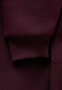 Burgundowa tkanina bluzy z prążkowanymi mankietami. Tekstura jest gładka z delikatnym połyskiem, prezentując bezszwowy projekt i subtelne detale.