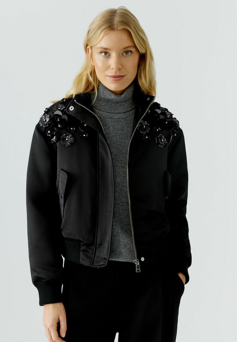 Schwarze Bomberjacke mit floralen Verzierungen an den Schultern, Reißverschluss und gerippten Bündchen, getragen über einem grauen Rollkragenpullover.