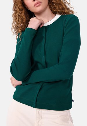 Frau mit lockigem Haar, die ein dunkelgrünes, langärmeliges Hemd mit Knöpfen über einem weißen Top trägt und ihren Hals mit einer Hand, die über den Körper gekreuzt ist, berührt.
