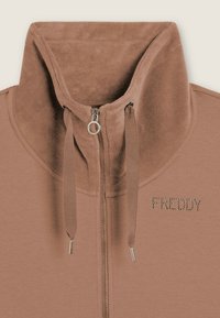 Sweat à zip marron avec un col haut, doté d'une texture lisse, d'un cordon de serrage et d'accents en cristal formant le mot "FREDDY" sur le devant.