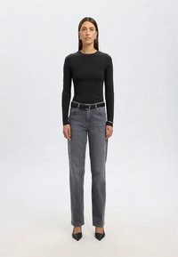 Kvinne står vendt fremover iført en svart langermet topp, grå høytsittende jeans med svart belte, og svarte spisse sko.