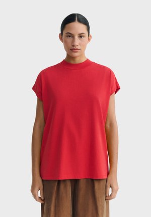 Femme aux cheveux foncés attachés, portant un haut rouge à manches courtes ample et une jupe marron, debout devant un fond gris clair uni.