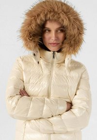 Chaqueta puffer beige con un acabado brillante, diseño de acolchado horizontal y capucha de piel sintética desmontable con un detalle en tono marrón.