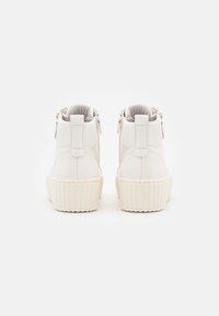 Para białych botków sięgających kostek z żebrowanymi podeszwami, tylnymi uchwytami i bocznymi zamkami, pokazanych z tyłu na białym tle.