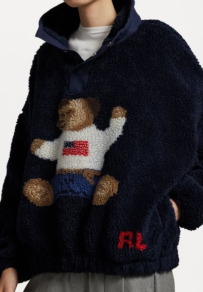 Marineblauer Fleece-Pullover mit hohem Kragen, der ein strukturiertes Teddy-Bär-Muster in Hellbraun und Weiß sowie ein rotes, gesticktes "RL"-Detail aufweist.