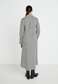 Prúžkovaný dlhý kabát s pásom v páse, vyrobený z ľahkej látky v čierno-bielych farbách, s dlhými rukávmi a rovnými strihom. Čierne členkové topánky.