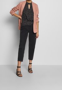 Donna che indossa un blazer rosa, una blusa nera a pois, jeans neri tagliati e sandali con tacco a strappo neri, in piedi contro un muro neutro.