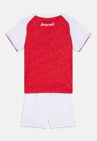 Röd tröja med ett texturerat mönster har vita ärmar och ordet "Arsenal" i vitt på baksidan; ihop med vita shorts.