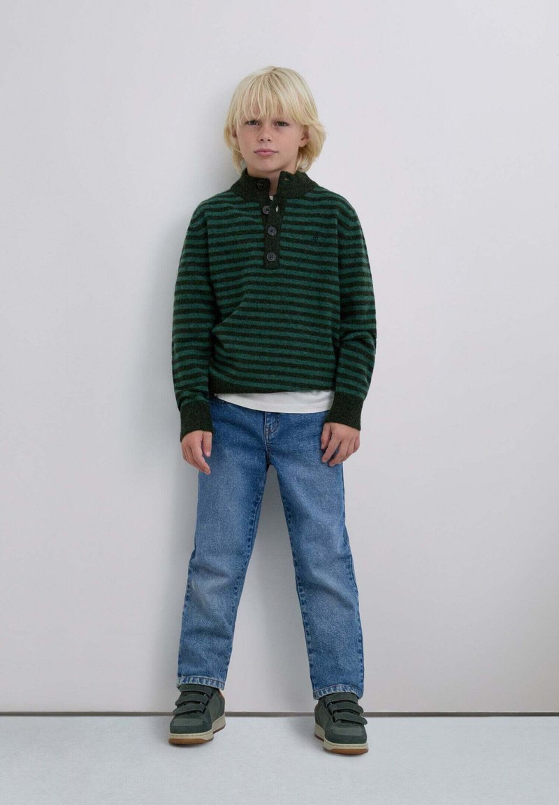 Suéter de rayas verdes y negras con cuello redondeado y tres botones, combinado con jeans azules y zapatillas verdes. Estilo sencillo y casual.