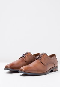 Braune Leder-Herrenschuhe mit glatter Oberfläche, runder Zehenform, blauen Akzenten an der Sohle und einem strukturierten Paneel nahe der Schnürsenkel.