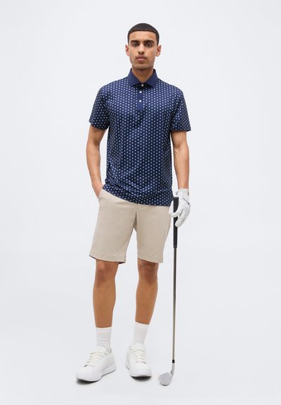Άνδρας με μπλε πουά πόλο, μπεζ σορτς, λευκά παπούτσια και γάντι, κρατώντας μπαστούνι γκολφ, στέκεται μπροστά σε λευκό φόντο.