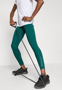 Žalios treniruočių tamprės su atspindinčiais akcentais, derinamos su balta sportine palaidine ir juodais sportbačiais, laikant pasipriešinimo juostą.