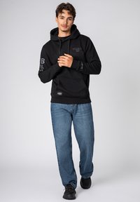 Zwarte hoodie met een voorzak, voorzien van bedrukte accenten op de mouwen. G gedragen met losse blauwe jeans en zwarte sneakers.