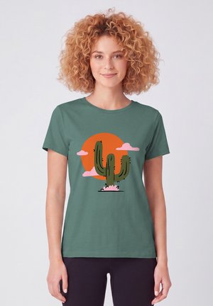 Jeune femme aux cheveux bouclés portant un t-shirt vert avec un cactus, des nuages roses et un soleil orange.